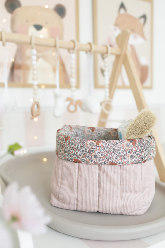 Das Bild zeigt ein selbstgenähtes Wickeltisch Utensilo aus rosa Cordstoff mit floralem Innenstoff. Die Aufbewahrung steht auf einem Wickeltisch mit Baby-Prints im Hintergrund