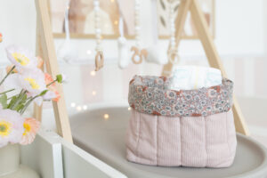 Das Bild zeigt ein selbstgenähtes Wickeltisch Utensilo aus rosa Cordstoff mit floralem Innenstoff. Die Aufbewahrung steht auf einem Wickeltisch mit Baby-Prints im Hintergrund